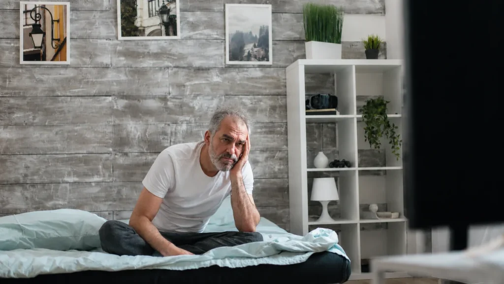 Man in his mid-60s sitting on the edge of his bed in early morning light with slumped posture, reflecting the unrefreshed waking that may signal sleep apnea or a circadian shift after 60.