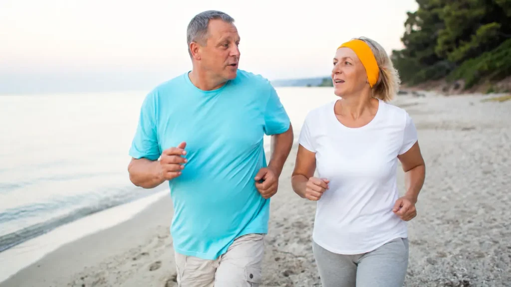 Two adults in their sixties jog side by side on a beach while mid-conversation, demonstrating the moderate-intensity aerobic exercise pace linked to cognitive health benefits.