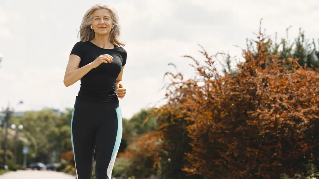 A woman over 50 jogging outdoors in a park with earbuds in, showing a calm and consistent daily exercise routine.