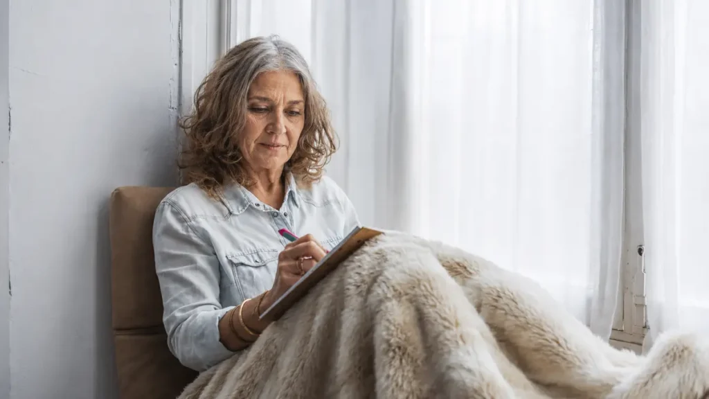 Older woman writing in a journal by a bright morning window wrapped in a blanket, tracking her sleep patterns and morning anxiety levels after 50.