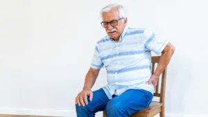 Mature man with glasses wincing in discomfort on a wooden chair while holding his lower hip to manage deep piriformis syndrome symptoms.
