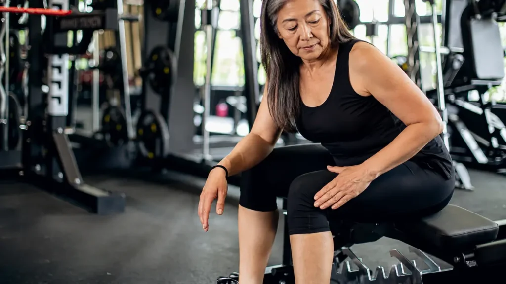 Mature East Asian woman checking her lower thigh muscle on a gym bench to evaluate normal physical feedback.