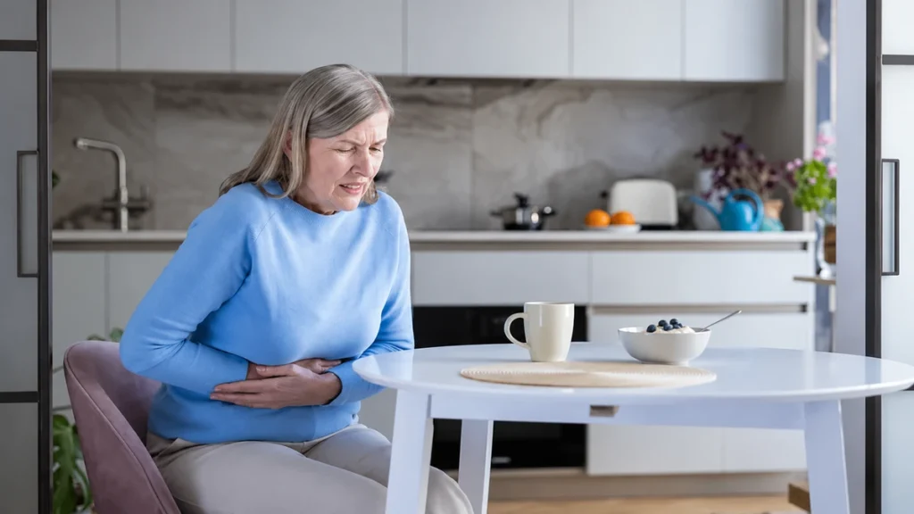 Mature woman with grey hair sitting at a kitchen table holding her stomach in discomfort next to a bowl of yogurt and a coffee mug.
