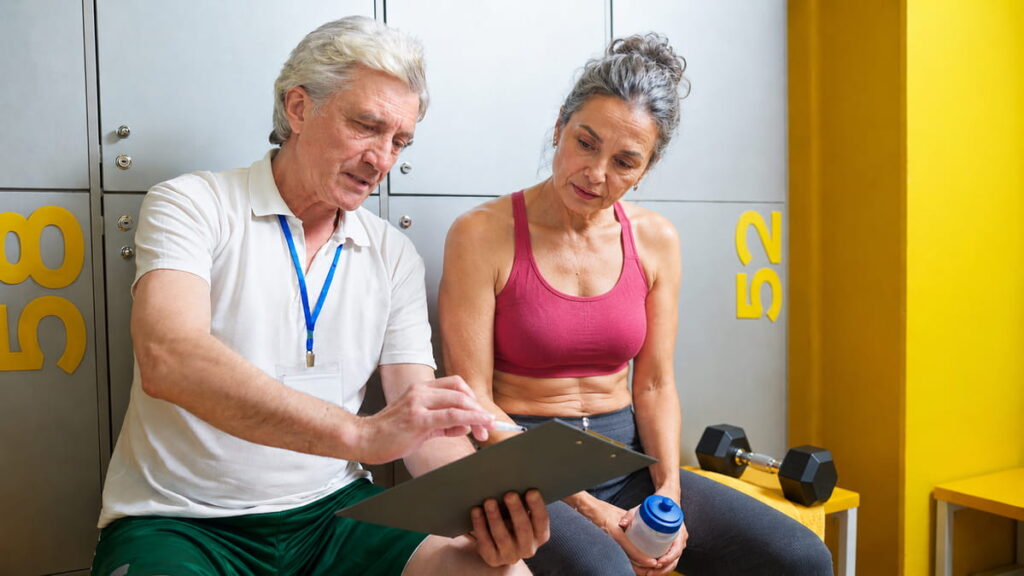 A mature woman reviewing training plan adjustments with her fitness coach on a locker room bench.
