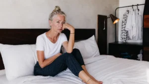 Mature woman with silver hair sitting on her bed looking exhausted, illustrating the reality of waking up tired after 8 hours of sleep due to age-related changes.