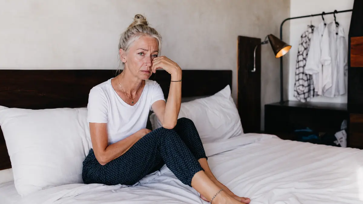Mature woman with silver hair sitting on her bed looking exhausted, illustrating the reality of waking up tired after 8 hours of sleep due to age-related changes.