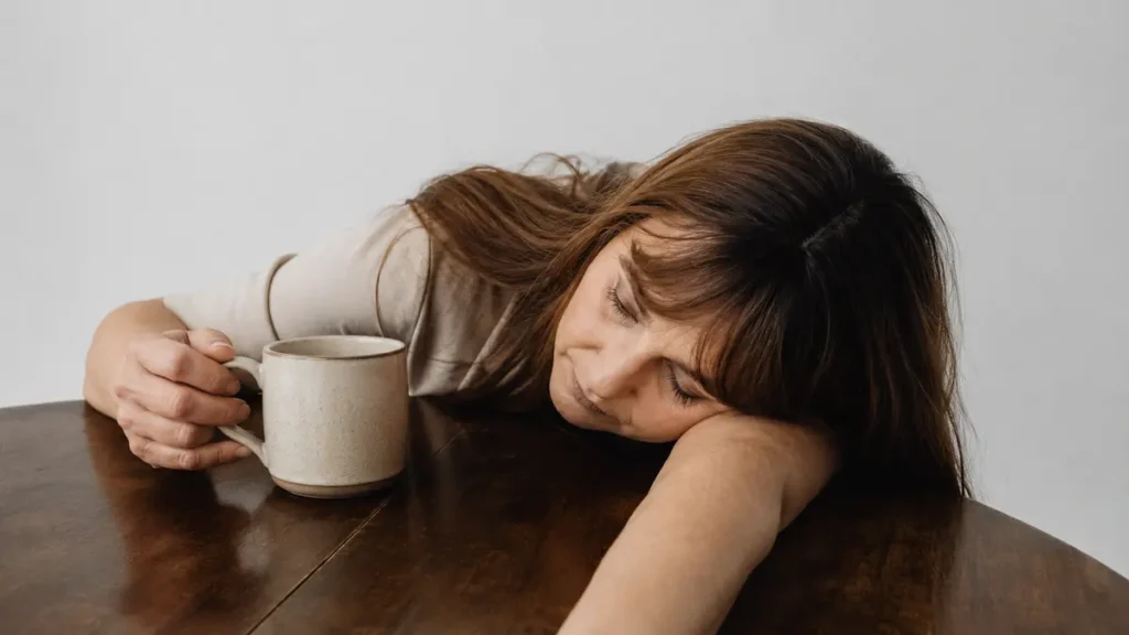 Exhausted midlife woman slumping over her kitchen table to cope with sudden perimenopausal mood instability and profound hormonal fatigue.