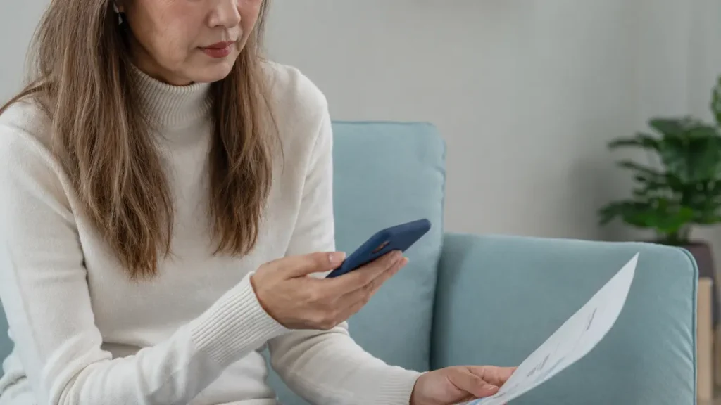 Focused midlife woman reviewing a printed medical document while tracking her cycle history on a smartphone app to prepare for her doctor visit.