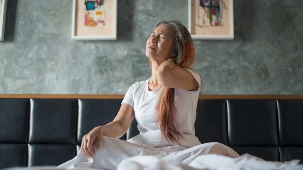 Mature woman sitting on bed holding her neck due to morning neck stiffness after 50.