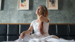 Mature woman sitting on bed holding her neck due to morning neck stiffness after 50.