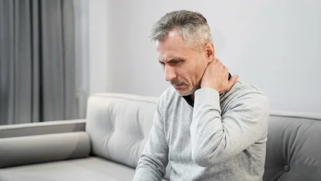Mature man sitting on a sofa holding the back of his neck with a concerned expression while experiencing prolonged morning neck stiffness.