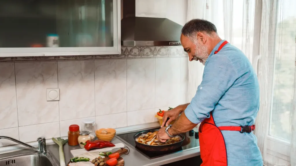 Mature man cooking chicken pieces in a frying pan alongside fresh chopped vegetables on a kitchen counter to prepare a whole-food meal that promotes a stable insulin release.