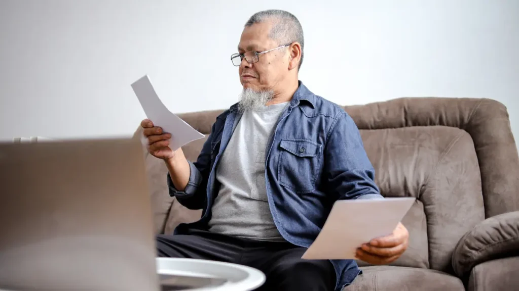 Mature man with a goatee reviews printed documents on a sofa to investigate hidden signs of impaired blood sugar control.