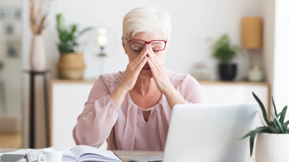 Mature woman pinching the bridge of her nose at her laptop, experiencing a sudden drop in afternoon focus.