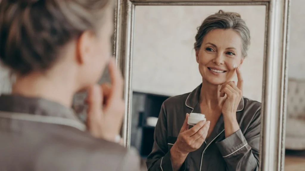 Mature woman in silk pajamas applies a dab of cream to her cheek while looking into a vanity mirror, protecting her skin from UV-induced matrix metalloproteinases that degrade deep dermal tissue.