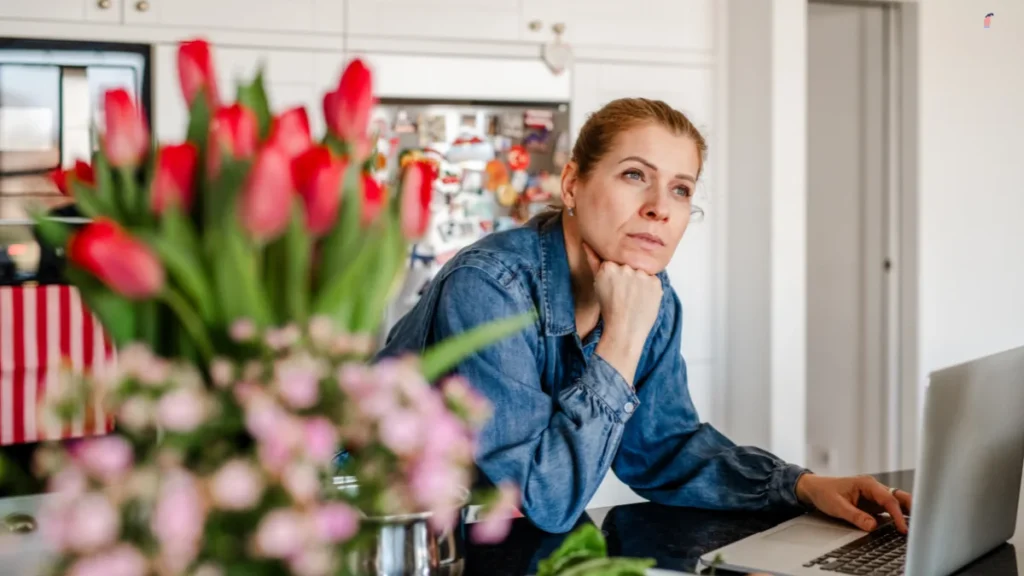 Pensive woman pausing at a kitchen laptop on a dark countertop to process feelings of social disconnection.