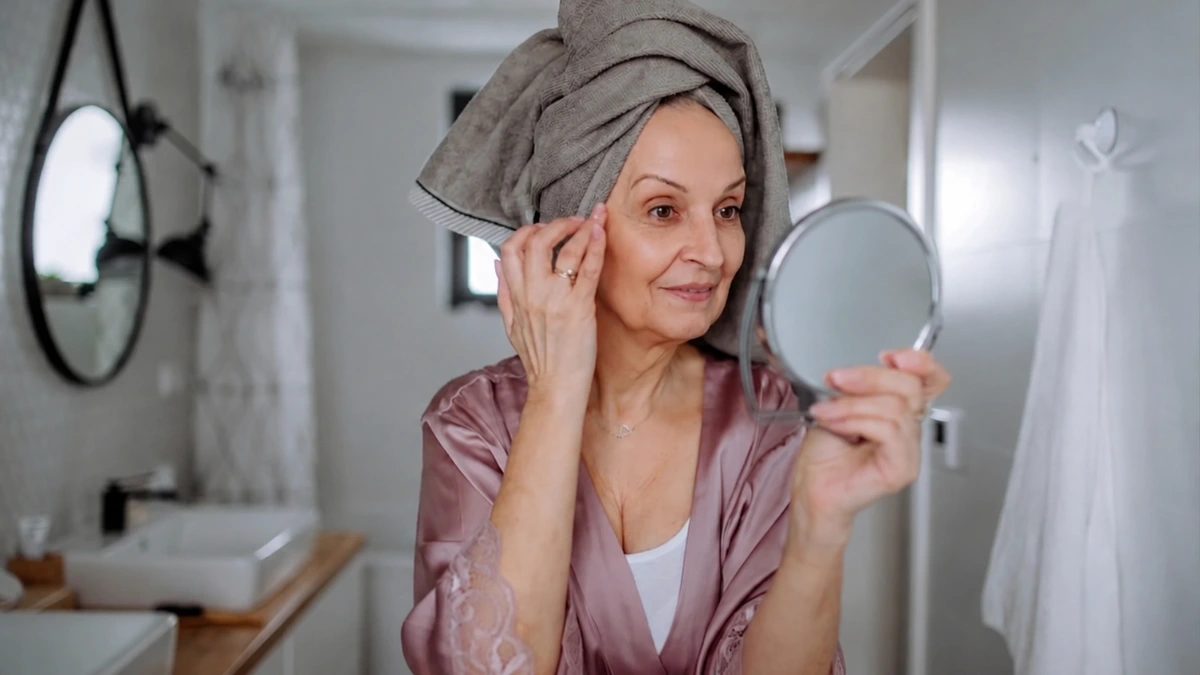 Mature woman with a towel wrapped around her hair looks into a handheld mirror while checking visible skin changes after 50 during her daily skincare routine, reflecting healthy aging and collagen support.