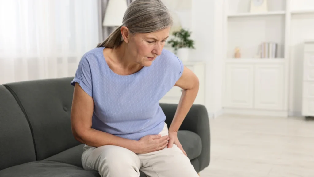 Woman in her 60s sitting on a sofa holding her side with a pained expression, experiencing lingering muscle soreness days after a workout.