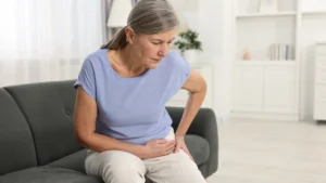 Woman in her 60s sitting on a sofa holding her side with a pained expression, experiencing lingering muscle soreness days after a workout.
