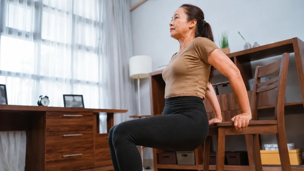Woman in her 50s gripping a sturdy chair seat while practicing a controlled sit-to-stand movement in a bright home living room.