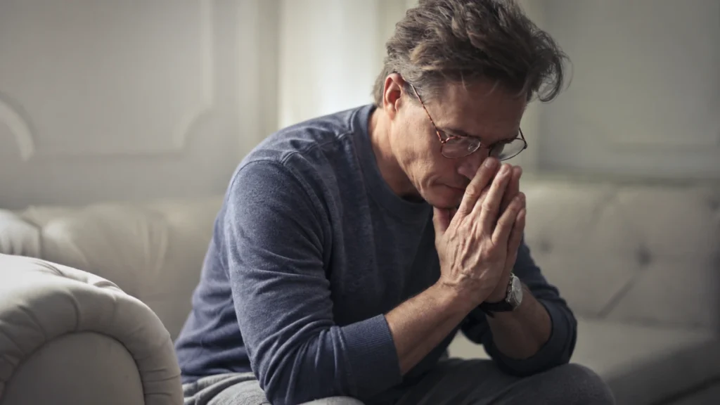 Man in his 50s sitting on a couch with head bowed and hands clasped, reflecting on gradual strength and muscle loss after 50.