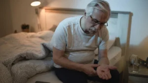 Mature man sitting up in bed at night, looking down with frustration at a small supplement pill in her open palm to manage sleep issues related to melatonin after 50.