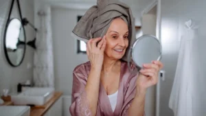 Mature woman with her hair wrapped in a towel smiles while looking into a handheld mirror, evaluating the structural shifts of her skin after 50 to track deep dermal preservation.