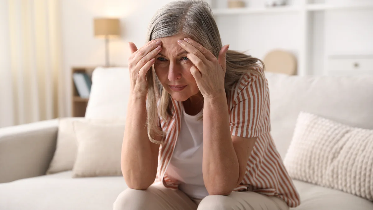 Mature woman rubbing her temples in a modern living room to clear sudden midday brain fog.