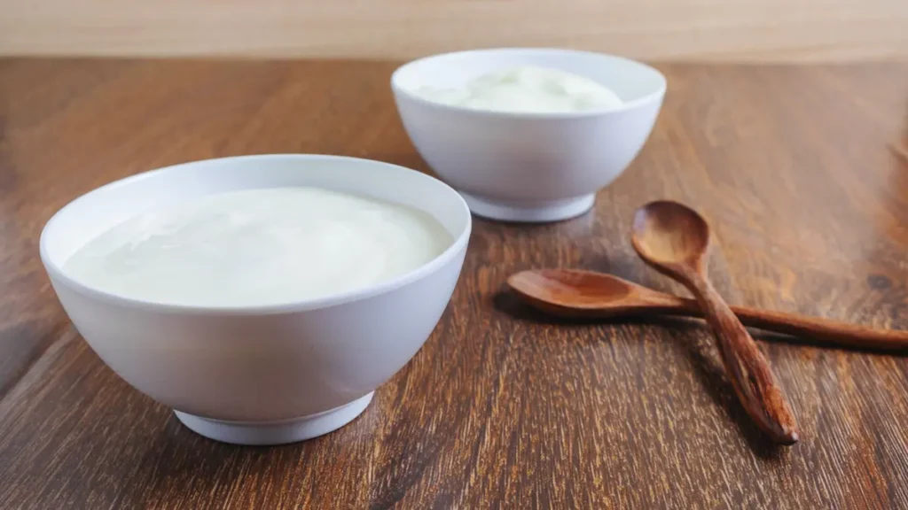 Two white bowls of plain yogurt with wooden spoons on a wooden surface representing lactose-free dairy alternatives.
