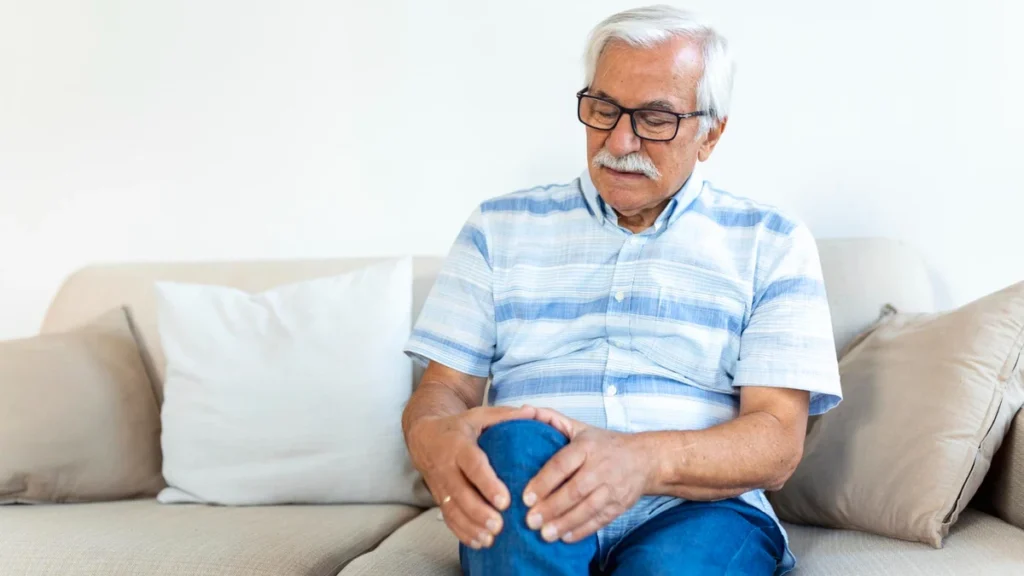 Older man seated on a sofa, both hands cupped around his knee, gaze fixed downward with a concerned expression as he examines it for swelling or tenderness.