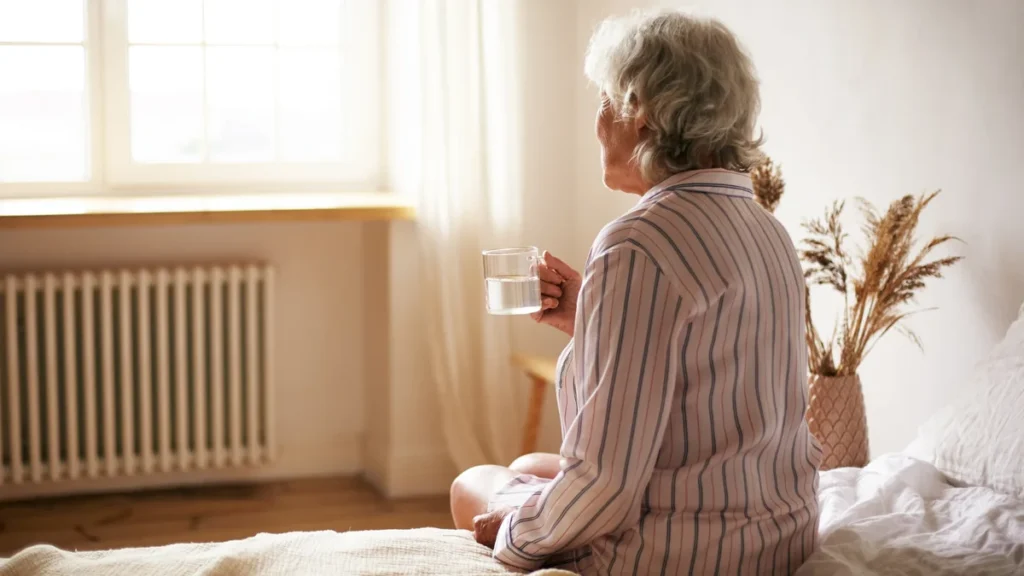 Mature woman sitting on a bed holding a glass of water, starting her morning with quiet reflection.
