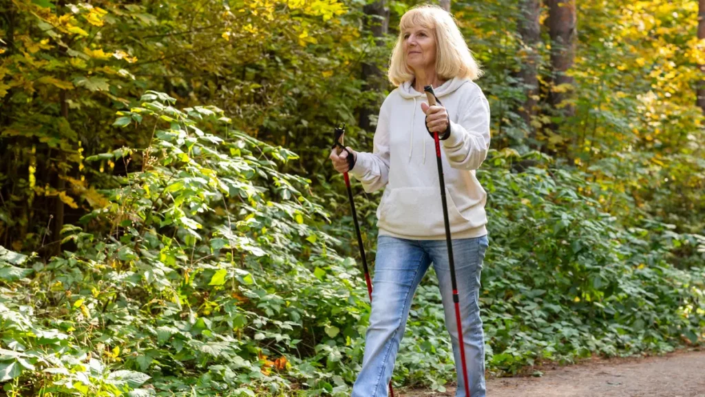 Active mature woman in a hoodie and jeans using walking poles on a sunny trail to protect her long-term physical trajectory.