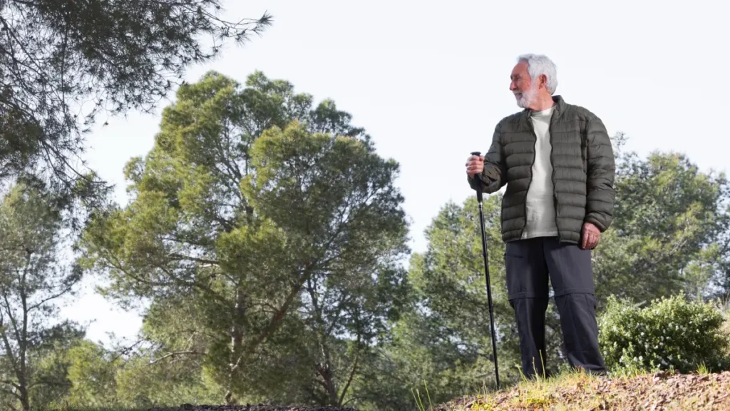 An active older man with a trekking pole hiking on a bright, tree-lined outdoor trail, maintaining his wellness routine even as seasonal sunlight and natural vitamin D synthesis change after sixty.