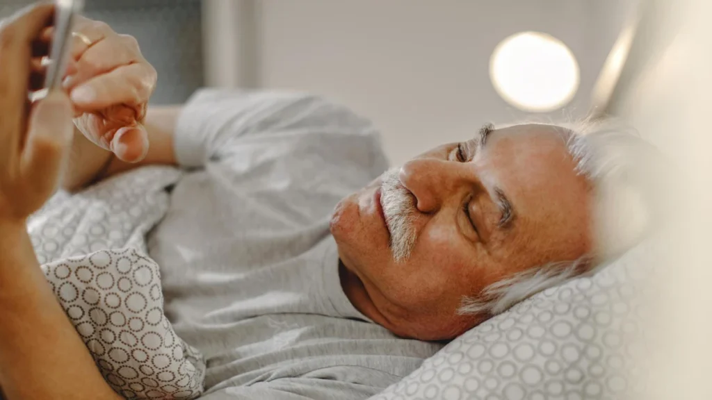 Tired mature man lying in bed looking at his phone under soft lamp light to track sleep fragmentation after waking up.
