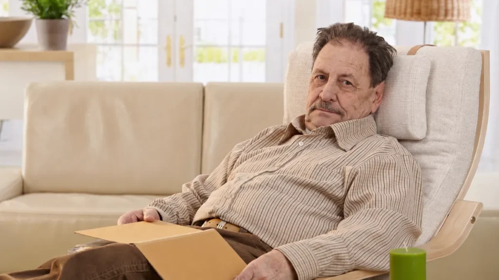 Weary older man in a striped shirt resting in a living room armchair with an open folder on his lap.