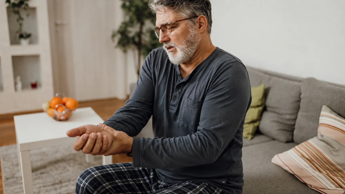 Thoughtful mature man in pajamas massaging his stiff hand while sitting on a living room sofa in the morning.