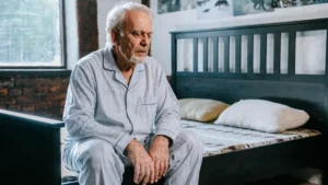 Mature man in blue pajamas reviews his physical wellness while sitting on a rustic bed after a vivid dream.