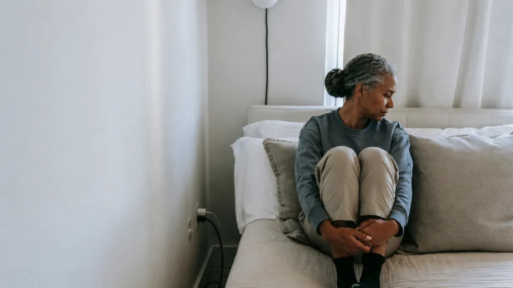 Exhausted mature woman sitting on bed hugging her knees, experiencing physical heaviness and flat mood of midlife depression.