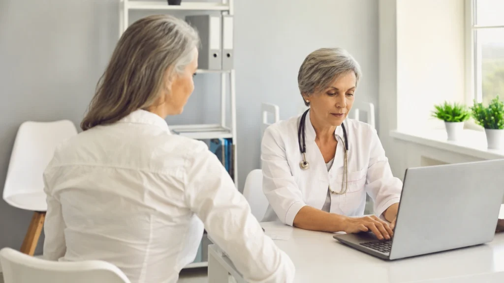 Mature female patient at a white desk waiting while her doctor types on a laptop during a medical consultation.