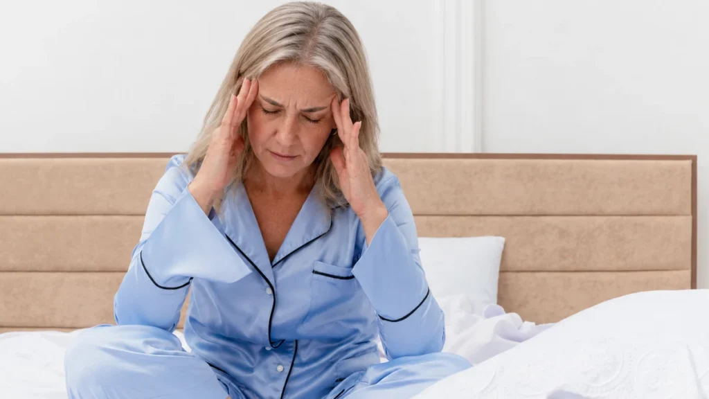 Tired mature woman in blue satin pajamas massaging her temples to soothe a waking headache from poor sleep.