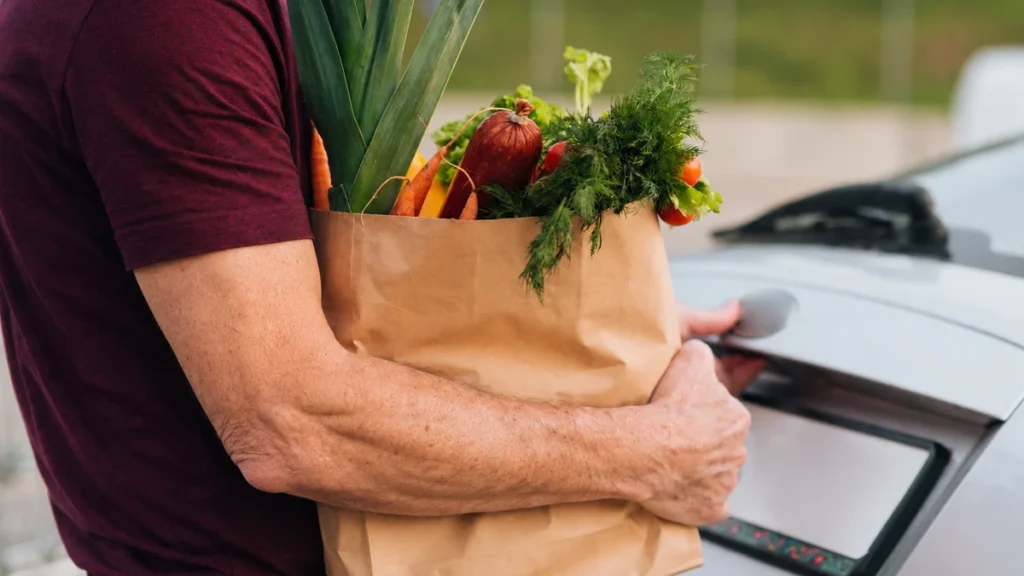 A mature man's arm gripping a paper grocery bag while opening a car trunk, highlighting daily functional strength.