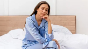 Mature woman sitting on a bed in morning light, looking downward to reflect on why she continues to wake up tired after 50.
