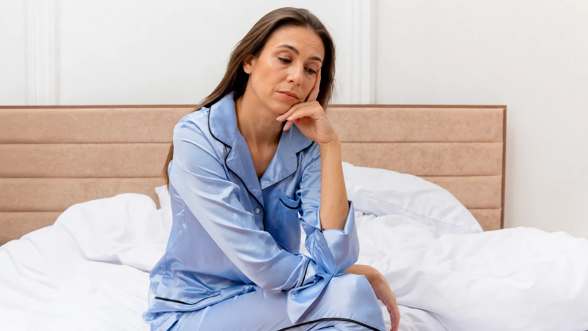 Mature woman sitting on a bed in morning light, looking downward to reflect on why she continues to wake up tired after 50.