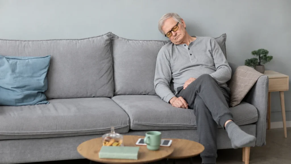 Senior man resting with closed eyes on a grey sofa to recover from overwhelming daytime exhaustion.