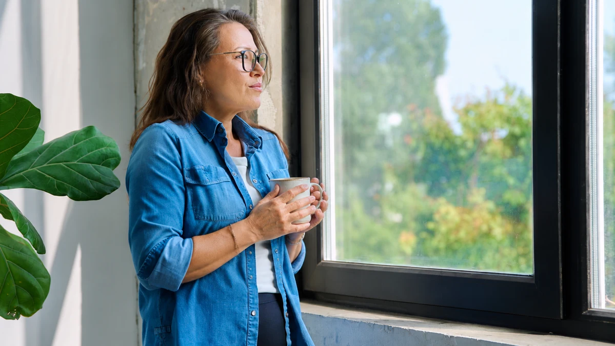 Woman in her 50s at window holding undrunk mug, gazing outward with preoccupied expression in survival mode after 50.