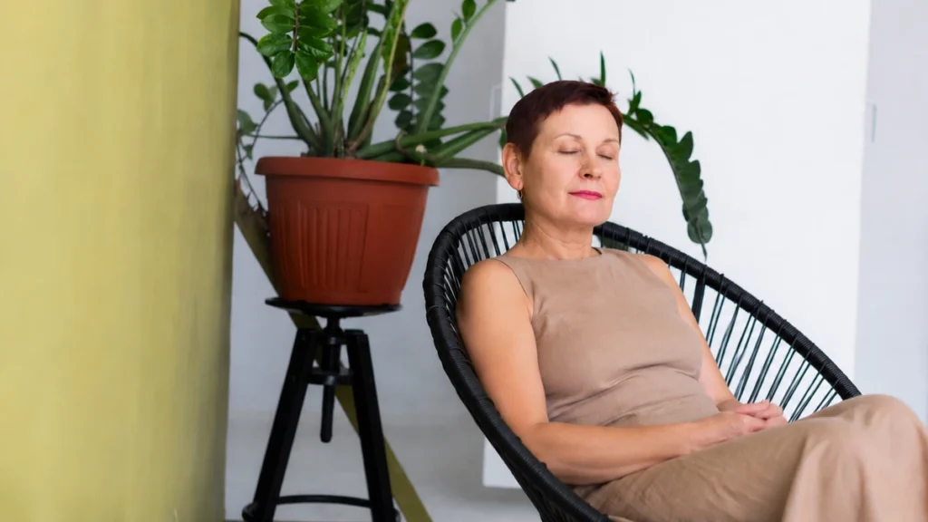 Woman in her 60s seated in rattan chair at home, eyes closed, hands in lap, shoulders released in deliberate breathing practice.