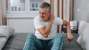 A mature man sitting on a bed wincing while rubbing his stiff shoulder in bright morning light, illustrating the sluggish physical recovery associated with age-related growth hormone release decline.