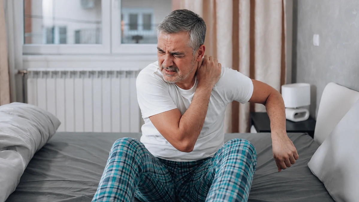 A mature man sitting on a bed wincing while rubbing his stiff shoulder in bright morning light, illustrating the sluggish physical recovery associated with age-related growth hormone release decline.