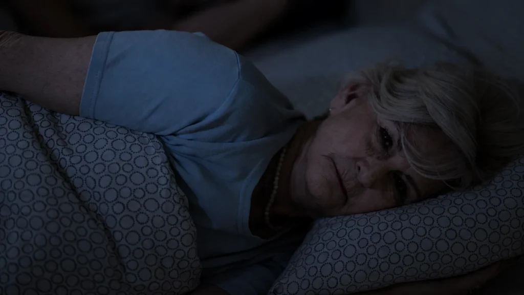 Mature woman lying awake in bed under a patterned blanket at night to manage age-related light sleep shifts.