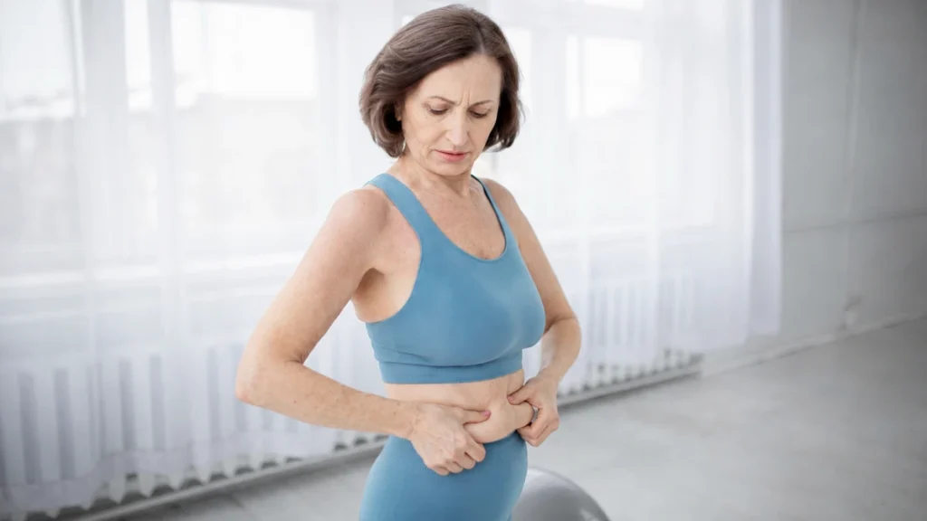 Mature woman in blue athletic wear looking down while pinching her midsection to track age-related metabolic shifts.